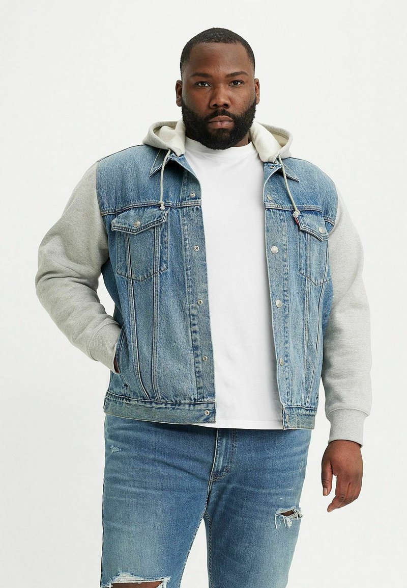 Chaqueta de mezclilla con mangas de sudadera gris, cierre frontal de botones y dos bolsillos en el pecho. Mezclilla azul clara con detalles desgastados y deslavados en los jeans.