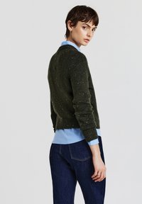 Dunkelgrüner gestrickter Pullover mit strukturierten Spritzern, Rundhalsausschnitt und gerippten Bündchen, über ein hellblaues Hemd mit Knopfleiste und dunklen Jeans getragen.