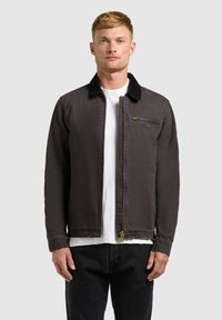 Homme portant une veste zippée marron foncé avec un col noir par-dessus une chemise blanche et un pantalon noir, debout devant un fond uni.