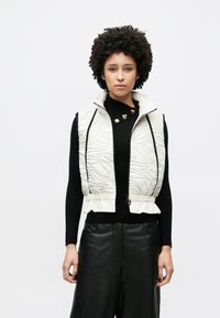 Steppweste in hellem Beige mit strukturiertem Muster, hohem Kragen, schwarzem Reißverschluss und gerafftem Saum. Trägt man über einem schwarzen Stricktop.