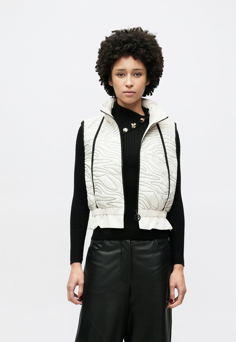 Gilet trapuntato beige chiaro con motivo testurizzato, collo alto, cerniera nera e orlo arricciato. Indossato sopra un top nero in maglia.