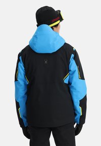 Giacca da sci nera e blu con dettagli gialli, dotata di cappuccio e logo a ragno sul retro. Realizzata in materiale impermeabile con una finitura texturizzata.