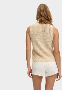 Top sin mangas de punto beige con bajo festoneado y patrón de tejido abierto, combinado con pantalones cortos de mezclilla blanca con bordes deshilachados.