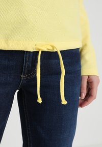 Gelber strukturierter Pullover mit Kordelzug am Saum, kombiniert mit dunkelblauen Jeans. Der Pullover hat ein locker sitzendes Design.