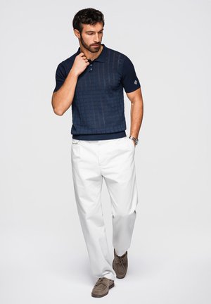 Man draagt marineblauw gebreid poloshirt, witte broek, bruine suède schoenen en polshorloge, staat met één hand in de zak en de andere dicht bij de kraag.