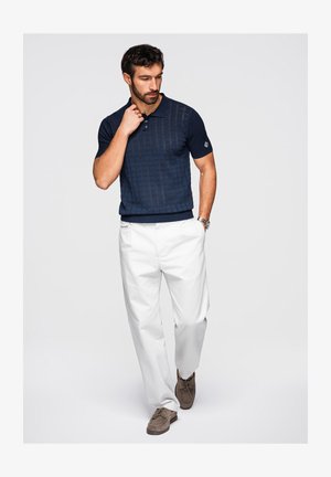 Mand iført marineblå strikket polo, hvide bukser, brune ruskindssko og armbåndsur, står med den ene hånd i lommen og den anden nær kraven.