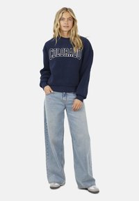 Marine crewneck sweatshirt met de tekst "COLORADO" in het wit, gecombineerd met lichtblauwe wijde jeans en grijze sneakers. Zachte textuur, casual pasvorm.