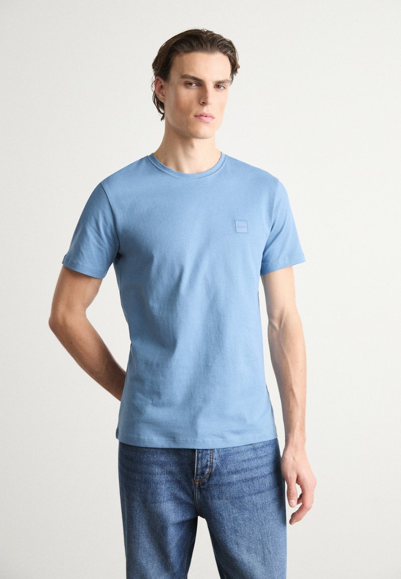 T-shirt de algodão azul claro com mangas curtas, gola redonda e uma pequena etiqueta no peito esquerdo. Combinado com jeans azul denim.