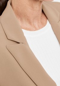 Blazer beige avec des revers nets, design structuré et texture lisse, associé à un haut blanc côtelé en dessous.