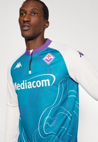 Vartininko marškinėliai žalsvai mėlynos ir baltos spalvų schemoje, abstraktiniu bangų raštu, purpuriniu apykakle ir akcentais, su ryškiai matomu „Mediacom“ logotipu.