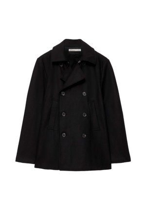 Personne portant une veste noire croisée, des lunettes de soleil foncées, un jean bleu et des chaussures noires, tenant un sac en cuir noir dans une main.