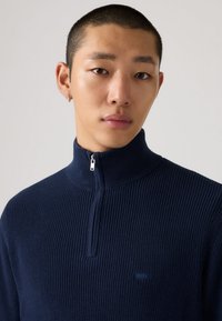 Tamsiai mėlynas megztinis su raižyta faktūra ir aukštu apykakliu su užtrauktuku. Pagamintas iš tekstūruotos megztos medžiagos, priekyje yra mažas, diskretiškas logotipas.