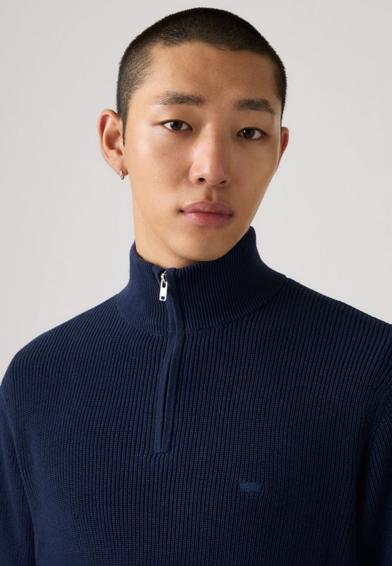 Marineblauer, gerippter Pullover mit hohem Reißverschluss-Kragen. Hergestellt aus einem strukturierten Strickstoff mit einem kleinen, dezenten Logo auf der Vorderseite.