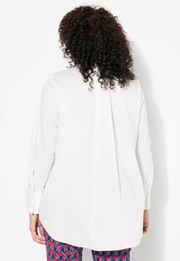 Blouse blanche à manches longues avec un pli dans le dos, des poignets boutonnés et un ourlet arrondi, associée à un pantalon coloré à motifs.