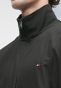 Άνδρας που φοράει μαύρο μπουφάν με ψηλό γιακά, μπροστινό φερμουάρ και μικρό κόκκινο, λευκό και μπλε λογότυπο στο στήθος.