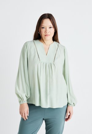 Blouse vert clair avec des manches longues bouffantes, un décolleté en V et des coutures décoratives le long du décolleté. Le matériau semble doux et fluide.