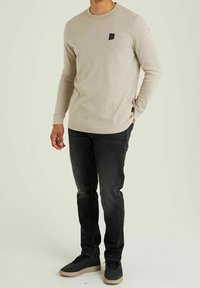 Light beige long-sleeve shirt with a small black logo on the chest, paired with black jeans and dark green sneakers. Smooth texture, fitted design.