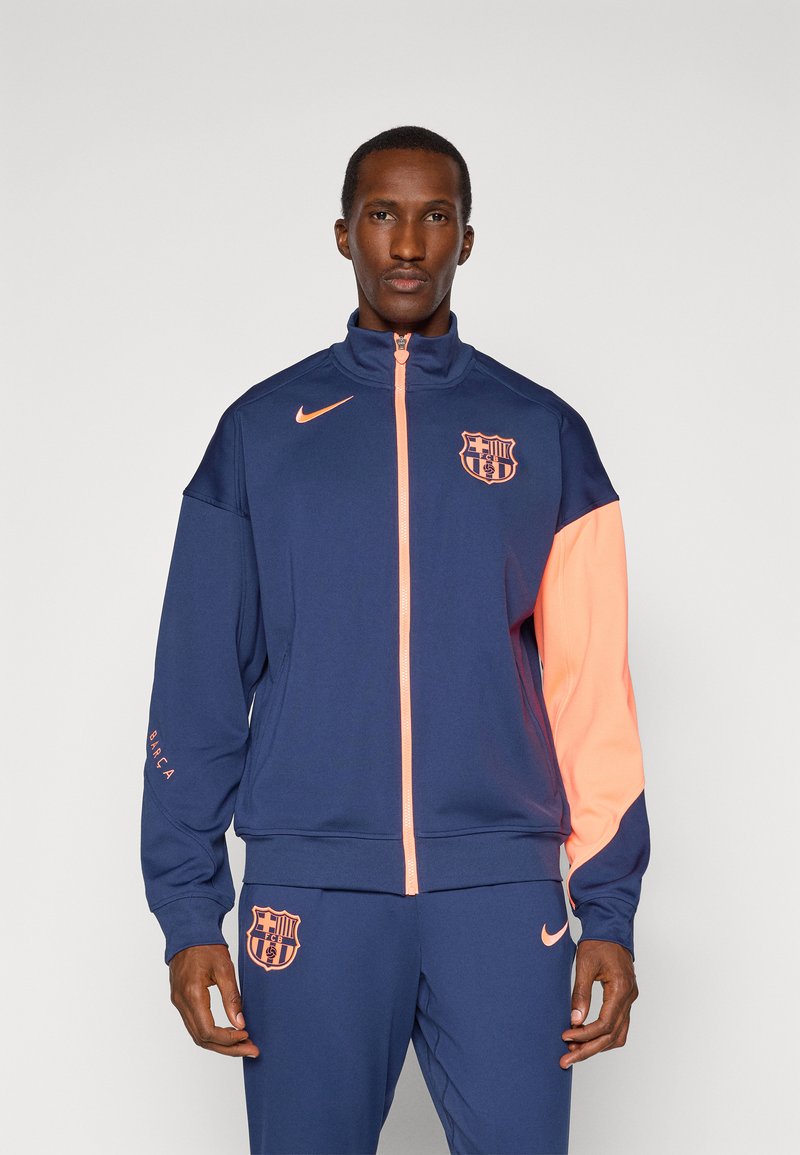 Nike Barcelona tracksuit, featuring a navy body, orange sleeve accents, logo on chest, and "BARCA" detail on cuff. Smooth texture.