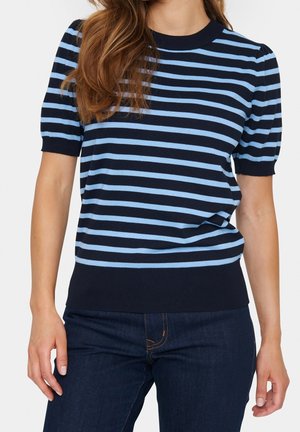 Kobieta w granatowo-niebieskiej bluzce w poziome paski z krótkim rękawem, w połączeniu z ciemnoniebieskimi jeansami, stoi z rękami swobodnie opuszczonymi wzdłuż ciała.