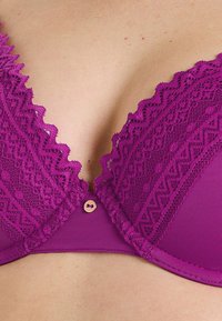 Soutien-gorge en dentelle violette avec bordures scallopées, un bouton décoratif au centre et une bande inférieure lisse. Dentelle texturée avec des motifs contrastants.