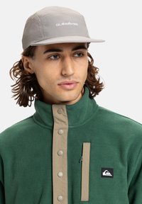 Cappello grigio con logo, abbinato a una giacca di pile verde con chiusura a bottoni e tasca con zip. Texture liscia, design a strati.