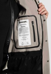 Gray inner lining of a jacket with a black mesh pocket, features a label with text, and a small zipper pocket on the side.