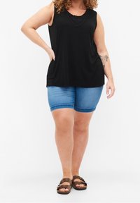 Kobieta w czarnej bezrękawnik z koronkowymi detalami, niebieskich szortach dżinsowych i brązowych sandałach na podwójnych paskach, stoi z ręką na biodrze.