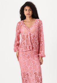 Blouse en dentelle rose avec des manches longues, décolleté en V et fermeture à boutons. Présente un motif floral et une texture transparente, assortie d'une jupe rose coordonnée.