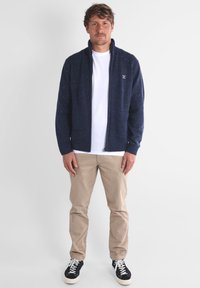 Un pull zippé bleu marine avec une finition texturée, des poignets et un col côtelés, porté par-dessus un t-shirt blanc, associé à un pantalon beige et des baskets noires.