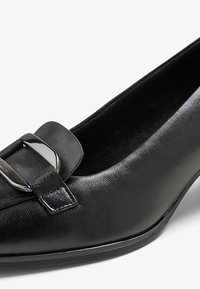 Escarpin en cuir noir à talon bas, doté d'un bout arrondi et d'une sangle ornée de métal à l'avant. Texture lisse avec une finition brillante.