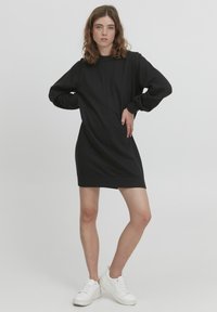 Abito felpa nero con maniche lunghe, colletto a costine e polsini aderenti. Texture liscia e silhouette a lunghezza ginocchio. Indossato con sneakers bianche.