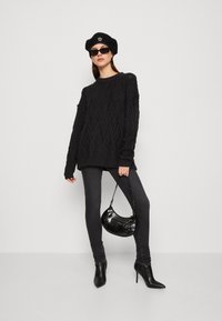 Μαύρο πλεκτό πουλόβερ με υφή σε σχέδιο διαμαντιού, συνδυασμένο με σκούρα στενά τζιν και μαύρες ψηλοτάκουνες μπότες σε κώνο. Τα αξεσουάρ περιλαμβάνουν μια μαύρη μπερέ και γυαλιά ηλίου.