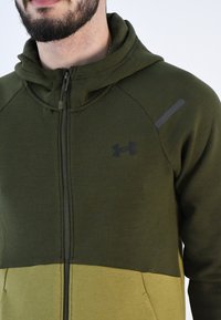 Sudadera con cremallera verde, con una sección en un verde más claro que contrasta, que cuenta con una capucha con doble forro, bolsillos con cremallera y un logo negro en el pecho.