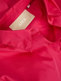 Czerwona satynowa koszula z wyższym kołnierzykiem, z metką "JJXX". Tekstura wydaje się gładka i błyszcząca, z guzikami jako detalami.