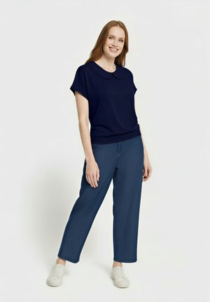 Lächelnde Frau mit langen roten Haaren, die ein marineblaues Kurzarmhemd mit Kragen, blaue, weit geschnittene Hosen und weiße Sneakers trägt, stehend vor einem weißen Hintergrund.