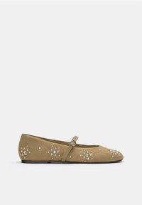 Chaussure plate en daim beige avec une lanière à boucle et des clous ronds argentés dispersés formant des motifs floraux sur les côtés et le bout.