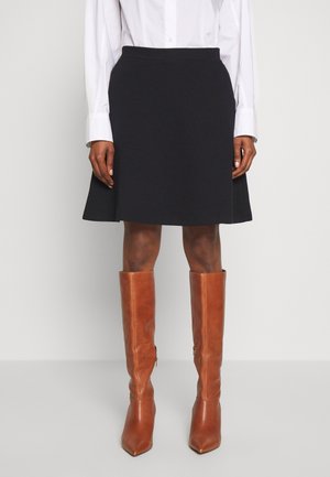 Person iført sort knælængde nederdel, hvid langærmet skjorte og høje lysebrune læderstøvler, stående mod en ensfarvet baggrund.