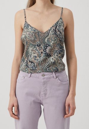 Kobieta w zielonej koszulce na cienkich ramiączkach w wzór paisley, wsuniętej w fioletowe spodnie z wysokim stanem i przednimi kieszeniami.