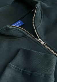 Ciemnozielona bluza z zamkiem błyskawicznym, z prążkowanym kołnierzem i mankietami, z widocznym metalowym zamkiem w kolorze srebrnym oraz etykietą marki umieszczoną wewnątrz kołnierza.
