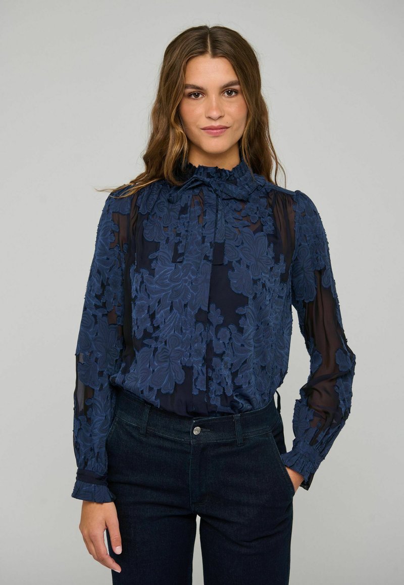 Femme portant un chemisier bleu marine transparent avec broderies florales et manches longues, rentré dans un jean sombre taille haute, debout avec une main dans la poche.