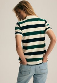 Polo w paski z krótkim rękawem w kolorach zielonym, granatowym i białym o teksturowanym wykończeniu. Zestawiony z jasnoniebieskimi dżinsami, prezentującymi luźny krój.