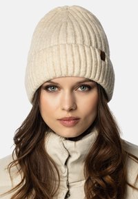 Crème gebreide beanie van textuurachtig garen, met een ribbelpatroon en een klein bruin leren label. Gecombineerd met een lichte jas.