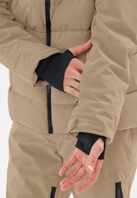 Beige isolierte Jacke mit schwarzen inneren Bündchen, sichtbarem Reißverschluss und Klettverschluss an den Ärmeln. Der Stoff wirkt glatt und gepolstert.