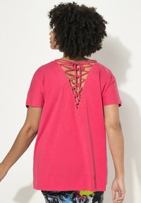 Rosa Baumwoll-T-Shirt mit V-förmigem Rückendesign und Schnürung, kurzen Ärmeln und lockerer Passform. Am Rücken leicht länger.