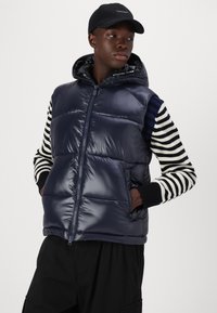 Vestă puffer navy cu închidere prin fermoar și glugă ajustabilă. Are mâneci lungi cu dungi alb-negru, cu accente ribbate.