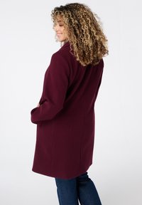 Manteau en laine bordeaux au silhouette ajustée, avec poches latérales, col cranté et texture lisse, associé à un jean en denim foncé.