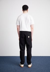 Muž s kudrnatými vlasy, oblečený v bílé tričko, černých kalhotách s nášivkou a bílých teniskách, stojící na modrém koberci čelem k bílé zdi.