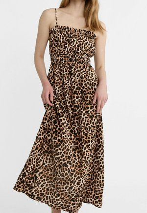 Vrouw draagt een mouwloze midi-jurk met luipaardprint, dunne bandjes en een geplooide taille, staand tegen een effen achtergrond.