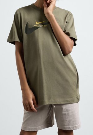 Person trägt olivgrünes Nike-T-Shirt mit schwarzen und gelben Swoosh-Logos und beigen Shorts, posiert mit einer Hand nahe am Gesicht.