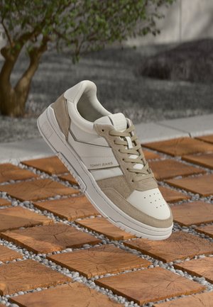 White and beige Tommy Jeans sneaker with laces, floating above a wooden tile and pebble outdoor floor with a tree in the background.